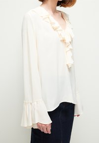 Frau trägt eine cremefarbene Bluse mit langen Ärmeln, Rüschenausschnitt und -manschetten, kombiniert mit einem dunkelblauen Jeansrock, vor einem schlichten hellen Hintergrund.