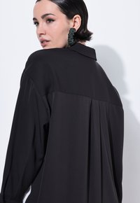 Schwarze Bluse mit lockerer Passform, langen Ärmeln und einem gefältelten Rückendesign. Sie hat einen Kragen und eine glatte, weiche Textur. Große grüne Ohrringe.