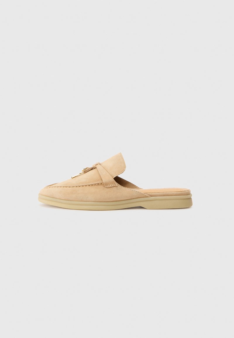 Beige suede slip-on shoe with a rounded toe, decorative stitching, and a gold-toned hardware accent. Flat sole and soft interior lining.