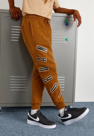 Person med bruna sweatpants med texten "JORDAN", svarta och vita Nike-sneakers, lutad mot ett grått metallskåp.