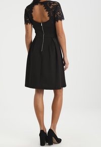 Vestito nero con maniche corte in pizzo, schiena scoperta e gonna plissettata. Presenta una chiusura con cerniera sul retro, abbinato a tacchi neri.