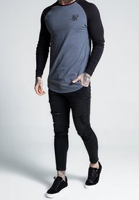 Chemise à manches longues grise avec des manches raglan noires et un logo brodé, associée à un jean skinny noir déchiré et des baskets noires.