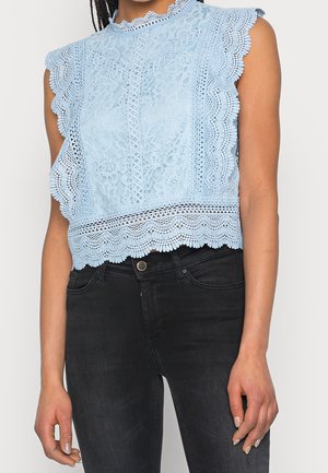 Ärmelloses hellblaues Spitzentop mit wellenförmigen Kanten, getragen zu hoch taillierten schwarzen Jeans, zeigt den Bauch und die Arme.