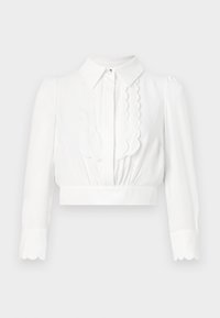Blusa bianca accorciata con colletto, dettagli scallop lungo la parte anteriore e maniche lunghe con polsini scallop. Tessuto leggero e liscio.