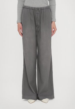Femme portant un pantalon large gris ample avec une taille à cordon, associé à un haut gris à manches longues et des chaussures blanches pointues.