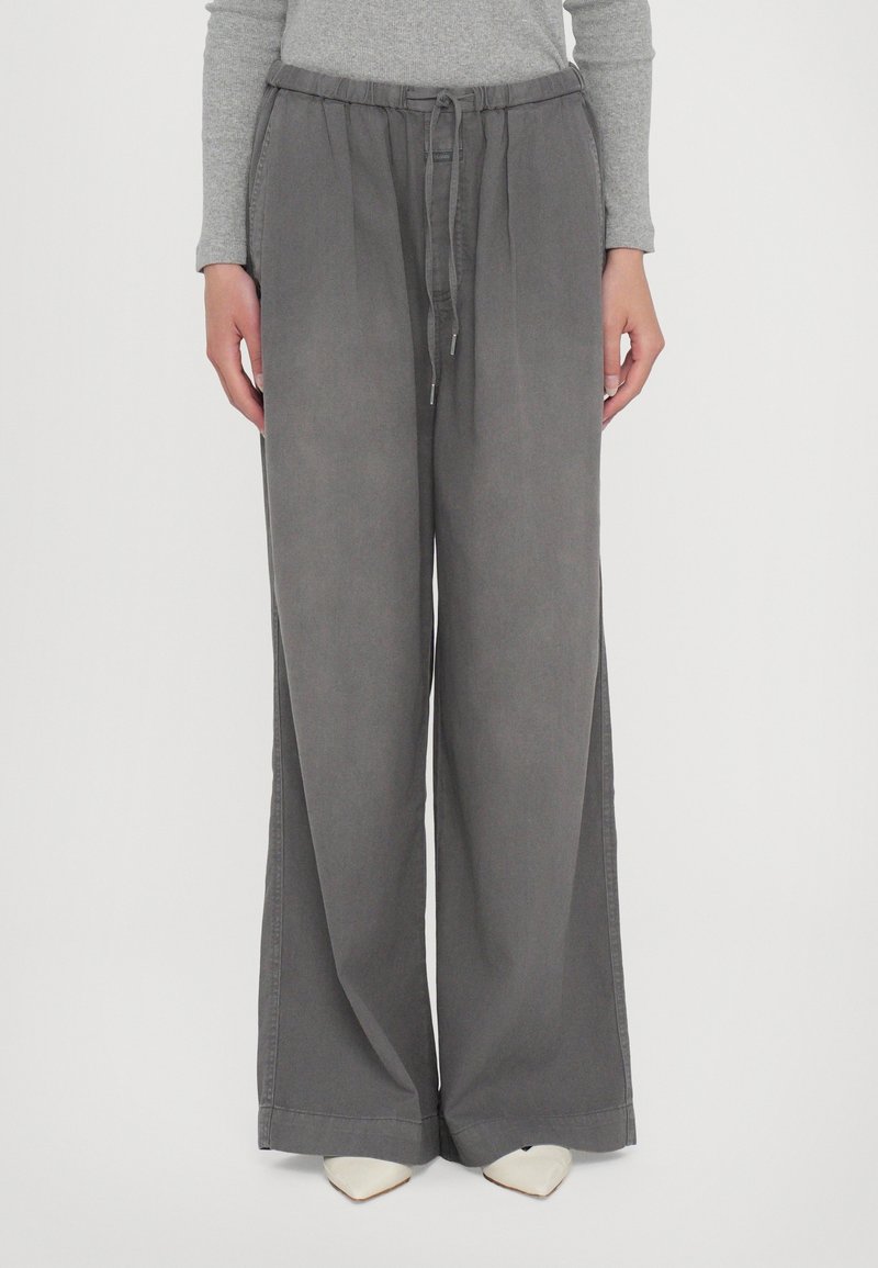 Femme portant un pantalon large gris ample avec une taille à cordon, associé à un haut gris à manches longues et des chaussures blanches pointues.