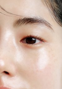 Primo piano di una pelle liscia e chiara con lentiggini visibili. L'occhio è marrone con ciglia sottili, e la pelle ha una texture lucida.