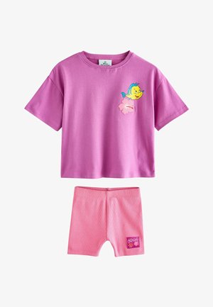 Niño pequeño con gorra rosa, camiseta morada con un dibujo de pez, pantalones cortos rosas, calcetines blancos y zapatillas rosas, de pie mirando hacia arriba.