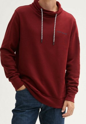 Person trägt einen dunkelroten Hoodie mit Kordeln und "Tom Tailor" Logo, kombiniert mit dunkelblauen Jeans, Hände in den Taschen.