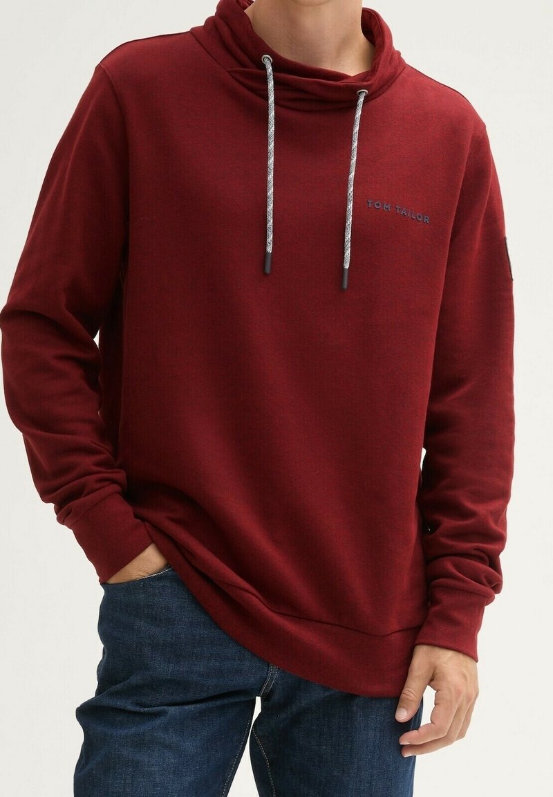 Personne portant un sweat à capuche rouge foncé avec cordons et logo « Tom Tailor », associé à un jean bleu foncé, mains dans les poches.