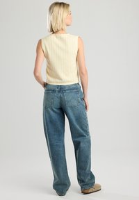 Light yellow knit sleeveless top with ribbed texture and scalloped neckline, paired with loose blue denim jeans featuring back pockets.