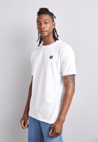 Vit bomullströja med korta ärmar, rund hals och en liten svart logotyp på vänster bröst. Modellen bär den med blå denimjeans.