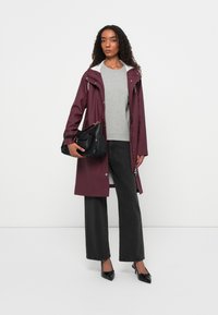 Casaco impermeável borgonha com capuz branco, suéter cinza e calças largas pretas. Mala preta com detalhes em dourado e sapatos de salto de bico fino.