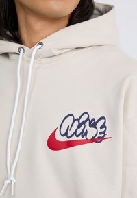 Et nærbillede af en person iført en beige hoodie med et stiliseret blåt og rødt Nike-logo på brystet, med hvide snore.