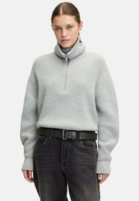 Hellgrauer Strickpullover mit hohem Kragen und halben Reißverschluss, mit weiten Ärmeln. Kombiniert mit dunkelgrauen Jeans und einem mit Nieten besetzten schwarzen Gürtel.