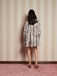 Žena s dlhými tmavými vlasmi stojí čelom k pruhovanej béžovej stene, oblečená v kvetinových šatách a leopardích podpätkoch na ružovom koberci.