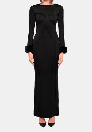 Woman wearing a fitted black long-sleeve floor-length dress with fur cuffs and pointed black heels, standing against a plain background.