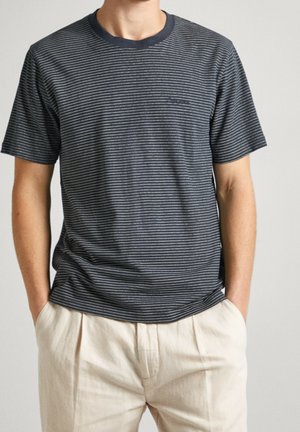 Mężczyzna ubrany w granatowy t-shirt z krótkim rękawem z cienkimi białymi poziomymi paskami oraz jasnobeżowe spodnie, ręce w kieszeniach.
