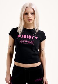 Fekete, rövidített póló, amelyen rózsaszín betűkkel szerepel a "JUICY" és az "Angel" felirat. Rövid ujjú, sima anyagból készült, karcsúsított fazonú. A karján tetoválások díszítik.