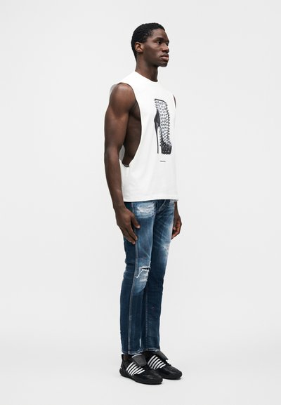 Λευκό αμάνικο μπλουζάκι με γραφικό σχέδιο, φθαρμένο μπλε τζιν και μαύρα παπούτσια με λευκές ρίγες. Πλάγια όψη του μοντέλου.