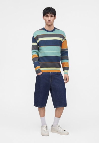 Jeune homme debout portant un pull rayé multicolore, un short en jean foncé jusqu'aux genoux, des chaussettes blanches et des baskets blanches, les mains dans les poches.