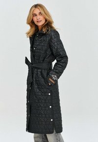 Czarny pikowany długi płaszcz z paskiem w talii, zatrzaskami i teksturowanym wzorem w diamenty. Zawiera dekoracyjną naszywkę na rękawie.