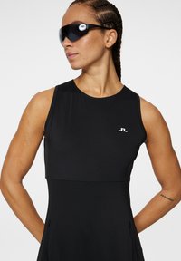 Robe de sport sans manches noire avec un corsage ajusté, texture lisse et détail de logo subtil sur le côté gauche. Comprend des lunettes de soleil sombres élégantes.