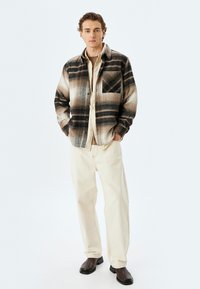 Camisa de franela a cuadros en beige y negro con un bolsillo en el pecho, combinada con pantalones crema y botas de tobillo marrones. Diseño simple y funcional.