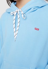 Jasnoniebieska bluza z kapturem wykonana z bawełnianej mieszanki, z przednią kieszenią, regulowanymi białymi sznurkami w czarne paski oraz czerwoną naszywką z logo.