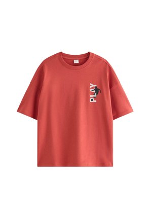 Dreng med brun beanie, rød t-shirt med teksten "PLAY", brune løse bukser, røde blomstrede sneakers og sølvkæde-halskæder, smilende med hænderne i lommerne.