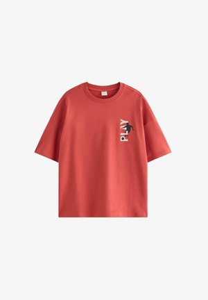 Dreng med brun beanie, rød t-shirt med teksten "PLAY", brune løse bukser, røde blomstrede sneakers og sølvkæde-halskæder, smilende med hænderne i lommerne.