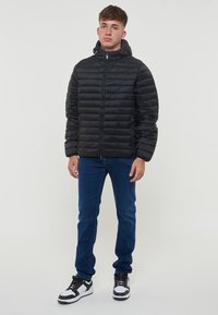 Giacca a vento nera con zip frontale, cappuccio e design trapuntato. Indossata con jeans blu e sneakers bianco e nere.