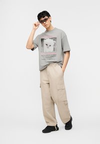 Young man wearing gray cat graphic t-shirt, beige cargo pants, black sneakers, and sunglasses, standing against a plain white background.