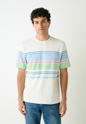 Jonge man met krullend haar draagt een wit kort mouwen shirt met blauwe, roze en groene horizontale strepen en blauwe spijkerbroek, staand tegen een witte muur.