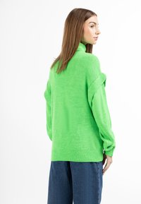 Pulover cu mânecă lungă și guler înalt, dintr-un material tricotat verde aprins, cu mansete și tiv cu dungi, purtat peste pantaloni din denim.
