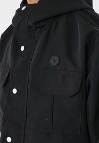 Giacca nera con cappuccio, caratterizzata da una finitura testurizzata, grandi tasche frontali e bottoni a pressione bianchi. Include un logo circolare sul petto.