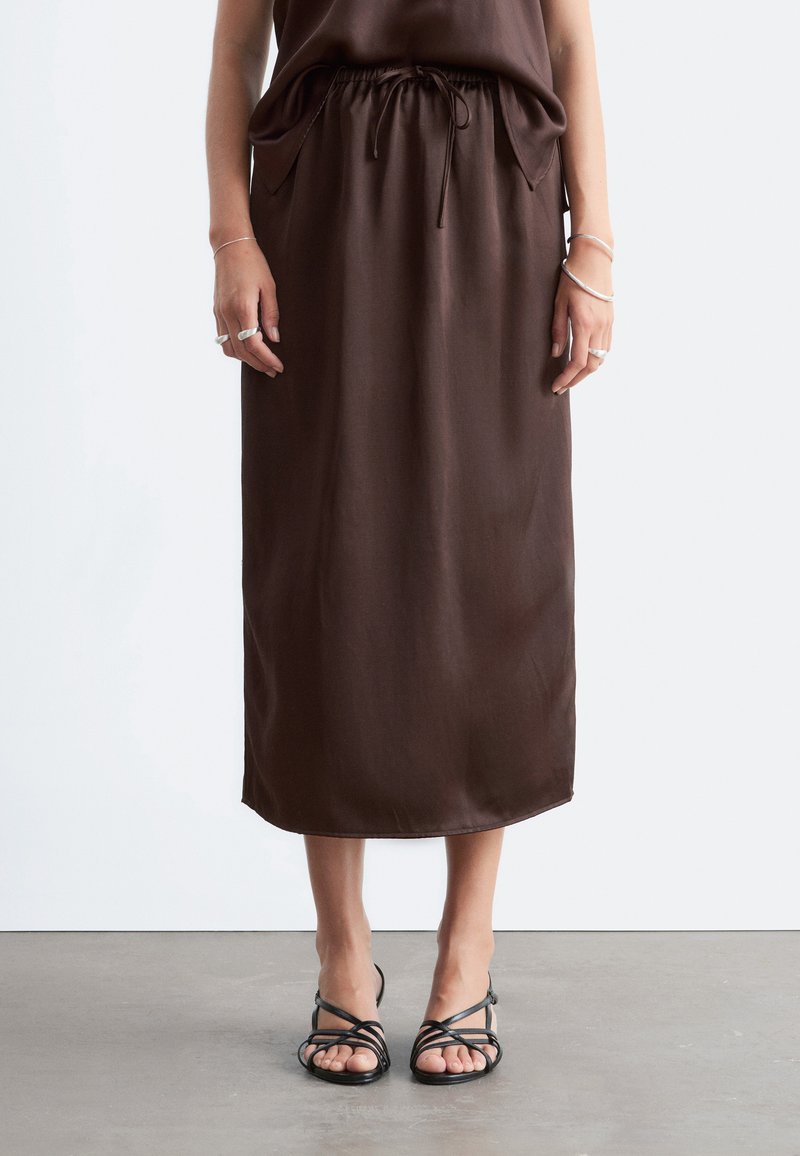 Personne portant une jupe midi marron avec une taille élastique et un cordon, associée à des sandales noires à brides et des bagues et bracelets argentés.