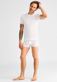 Hvit bomulls T-skjorte og matchende boxershorts, tettsittende design, korte ermer, glatt tekstur, båret av en person med lys hud og tatoveringer.
