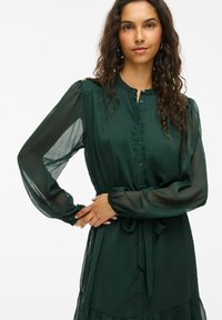 Rochie verde închis, cu nasturi, cu mâneci lungi din material transparent și talie strânsă. Dispune de un materialtexturat și un design cu fuste în straturi.