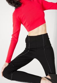 Vrouw in een felrode cropped langemouwen top en hoge zwarte broek met witte stikdetails, poseert tegen een effen achtergrond.