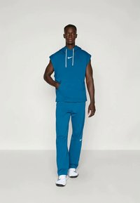 Άνδρας στέκεται φορώντας μπλε αμάνικο φούτερ Nike, ταιριαστούς μπλε παντελόνες και λευκά αθλητικά παπούτσια Nike, μπροστά σε απλό λευκό φόντο.