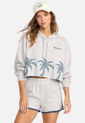Vrouw in een lichtgrijze cropped hoodie met palmbomenprints, bijpassende shorts met marineblauwe rand, en een witte pet met een surflogo.