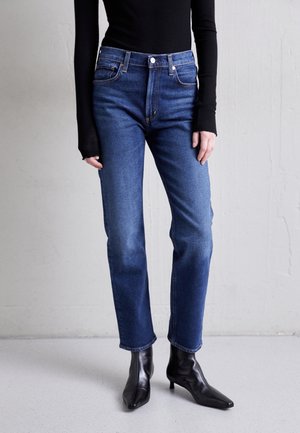 Persona vestida con jeans azules de pierna recta, top negro ajustado de manga larga y botas negras hasta el tobillo, de pie contra una pared lisa y clara.