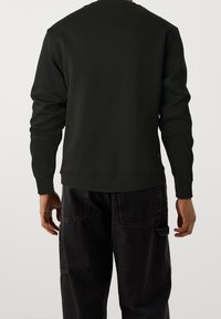 Sweatshirt nero a maniche lunghe con colletto tondo, con polsini e orlo a coste. Presenta una piccola toppa sul retro. Abbinato a pantaloni cargo scuri.