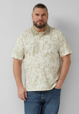 Mann mit Bart, der ein beiges, gemustertes Poloshirt und blaue Jeans trägt, steht mit einer Hand in der Hosentasche vor einem einfarbigen Hintergrund.