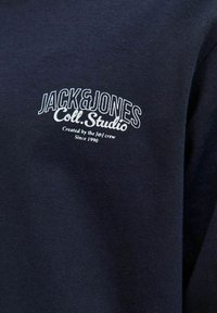 Tamno plava dukserica s mekanom teksturom, koja sadrži bijelu izvezenu oznaku: "JACK & JONES Coll. Studio" i "Kreirano od strane Je-I ekipe od 1990."