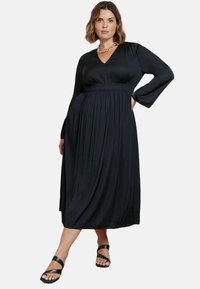 Svart plisserad klänning med långa ärmar, V-ringning och elastisk midja; matchad med svarta sandaler med remmar. Slät tygtextur.