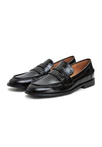 Paire de mocassins noirs en cuir à enfiler avec finition texturée et bride penny, doublure intérieure beige, et petits talons plats sur fond blanc.