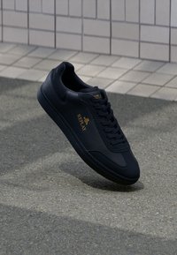 Schwarze Leder Sneaker mit strukturierter Oberfläche, runder Zehenpartie und flachen Schnürsenkeln. Mit einem goldenen Logo und einer Gummisohle mit strukturiertem Muster.