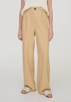 Model draagt beige wijde broek met plooien aan de voorkant, een zwarte knoop en witte open-toe sandalen tegen een effen achtergrond.